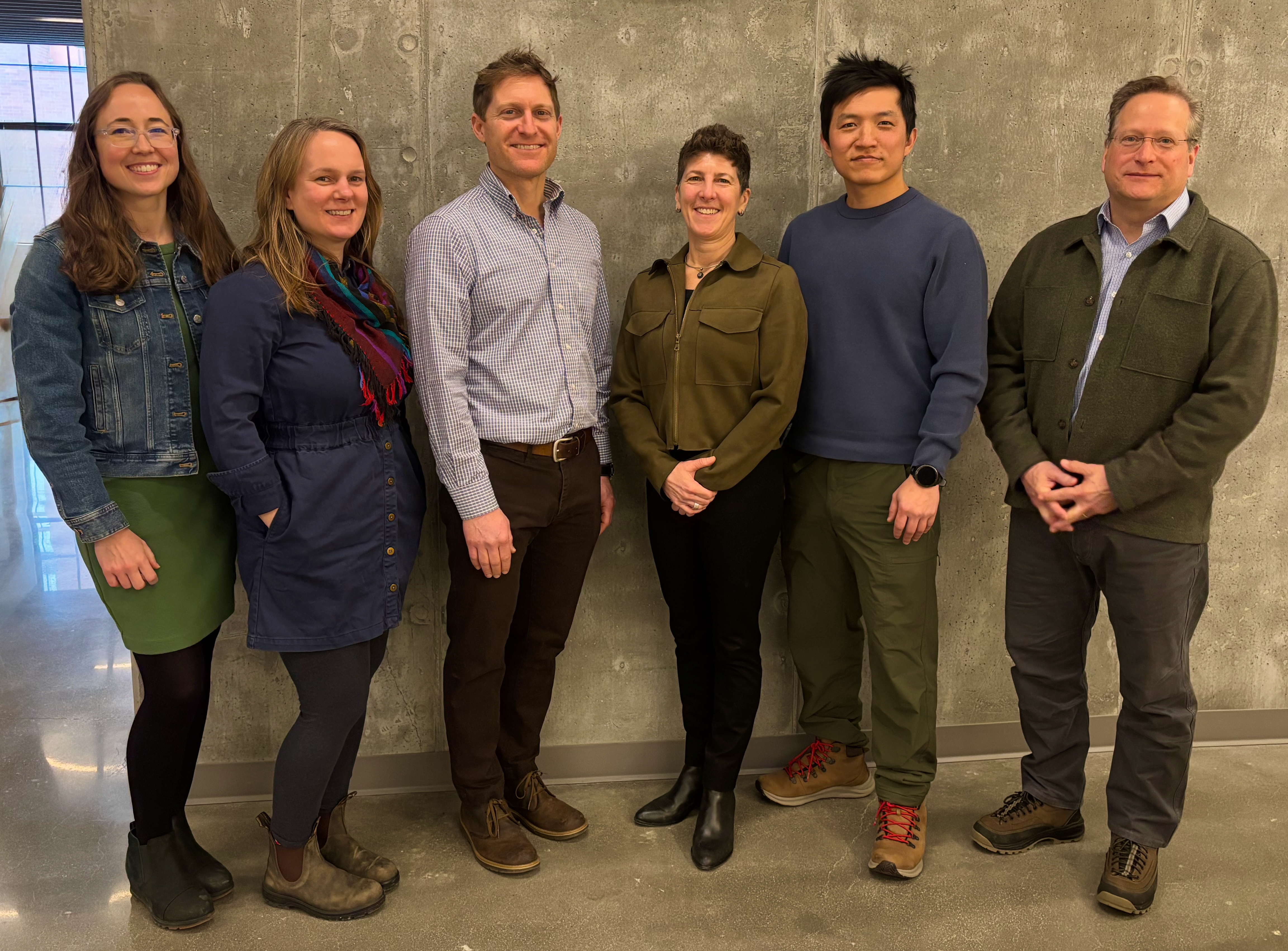 Six people stand smiling in front of a gray wall. They wear casual attire, expressing a friendly and relaxed atmosphere.