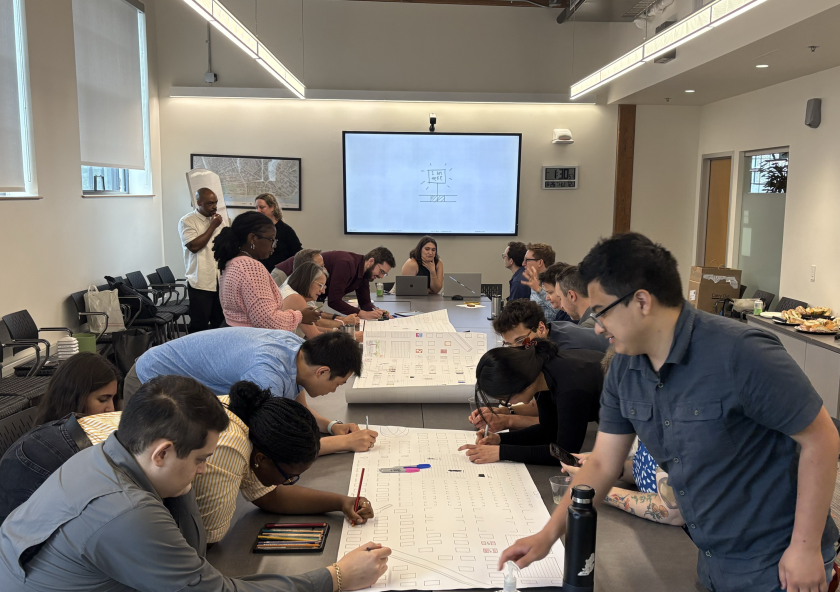 A group of MIT students and staff gathered around a conference table are actively engaged in a collaborative activity, writing on large sheets of paper. The room is well-lit, with a presentation screen in the background. The atmosphere is focused and collaborative.