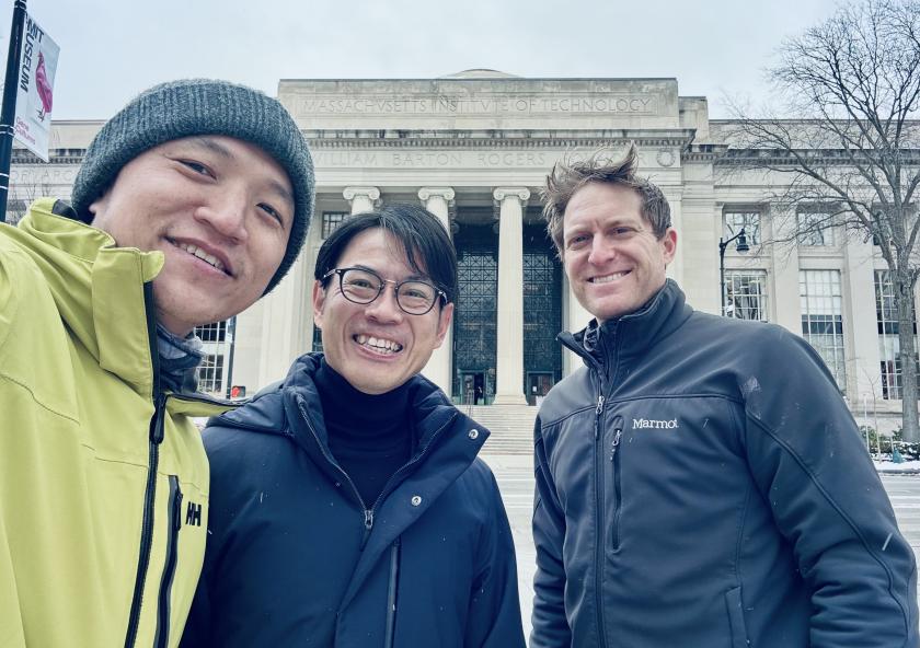 Three people smiling in front of a grand, columned building with "Massachusetts Institute of Technology" inscribed above the entrance. They appear cheerful and bundled warmly, suggesting a cold day.