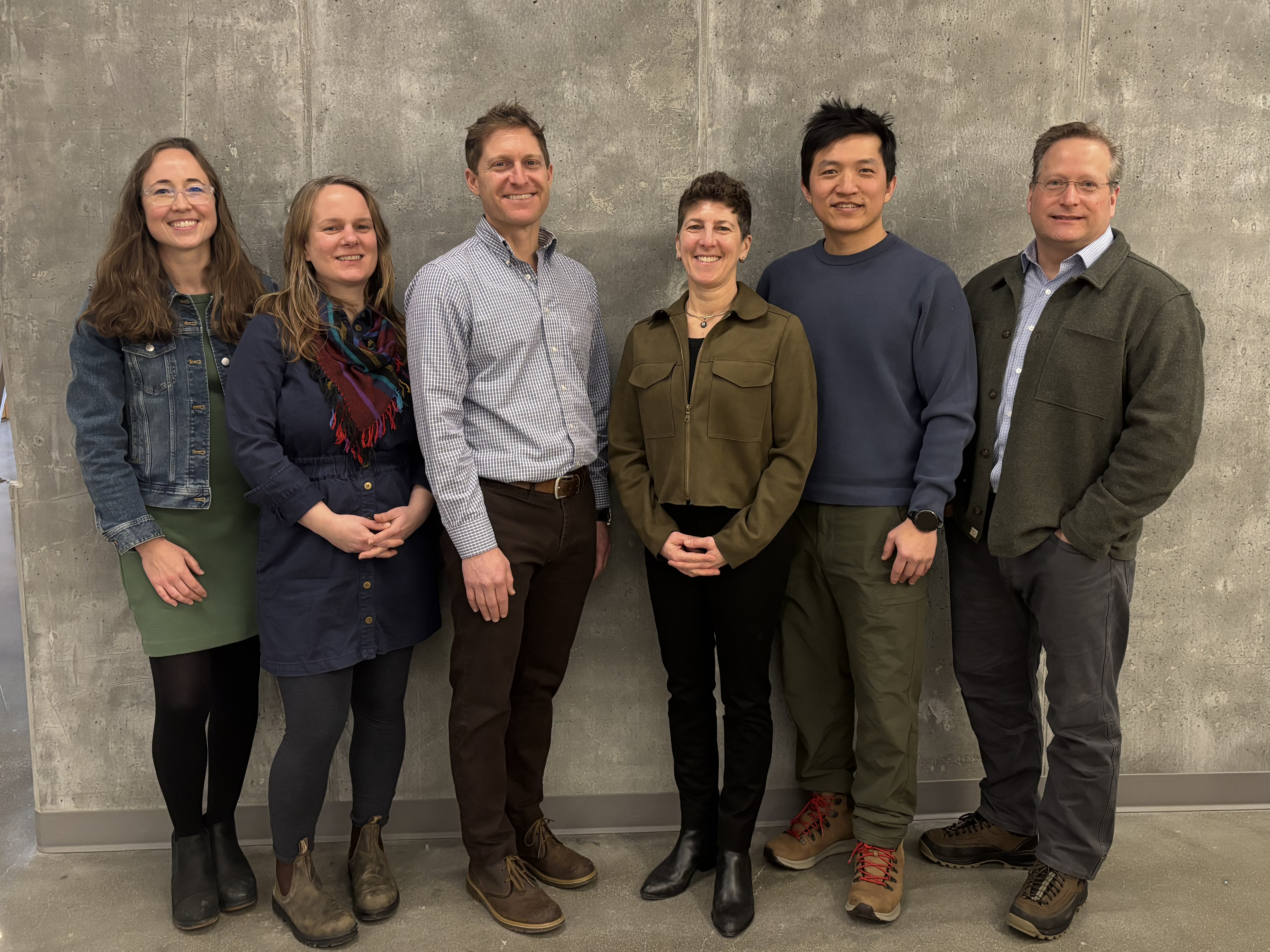 Six people stand smiling in front of a gray wall. They wear casual attire, expressing a friendly and relaxed atmosphere.