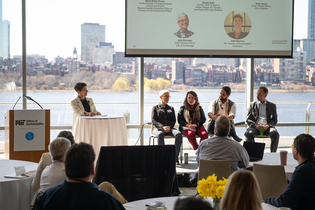 A panel discussion at the Sustainability Connect event with four people seated on stage and a moderator at a podium. Large windows show a cityscape and river view. Attendees sit at round tables, listening intently. The atmosphere is engaged and professional.