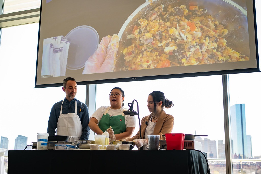 Three people in a cooking demonstration with a large screen showing a close-up of a colorful dish. The atmosphere is collaborative and engaging.