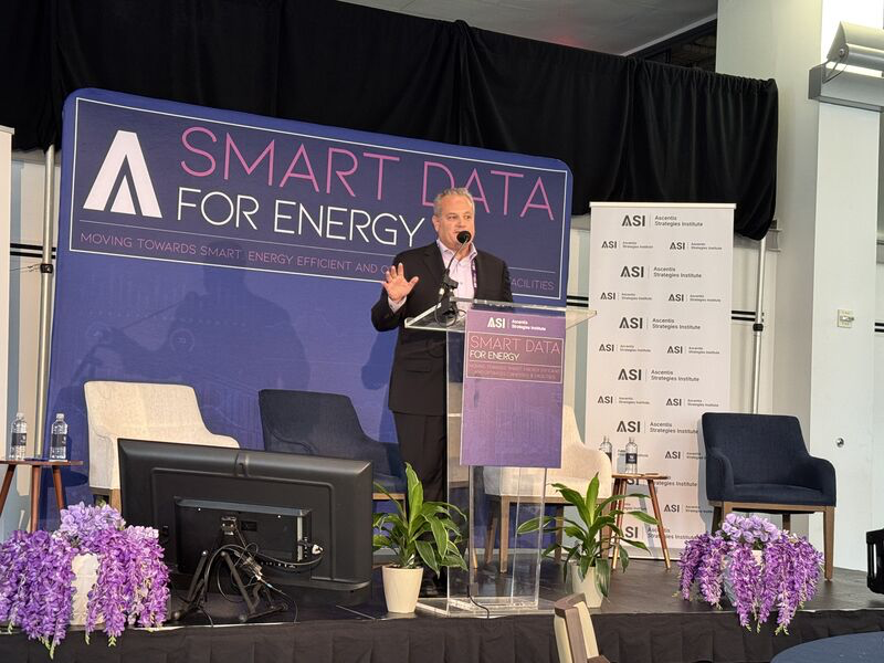 A man speaks at a podium on a stage with purple flowers and plants. The backdrop and podium read ASI "Smart Data for Energy"