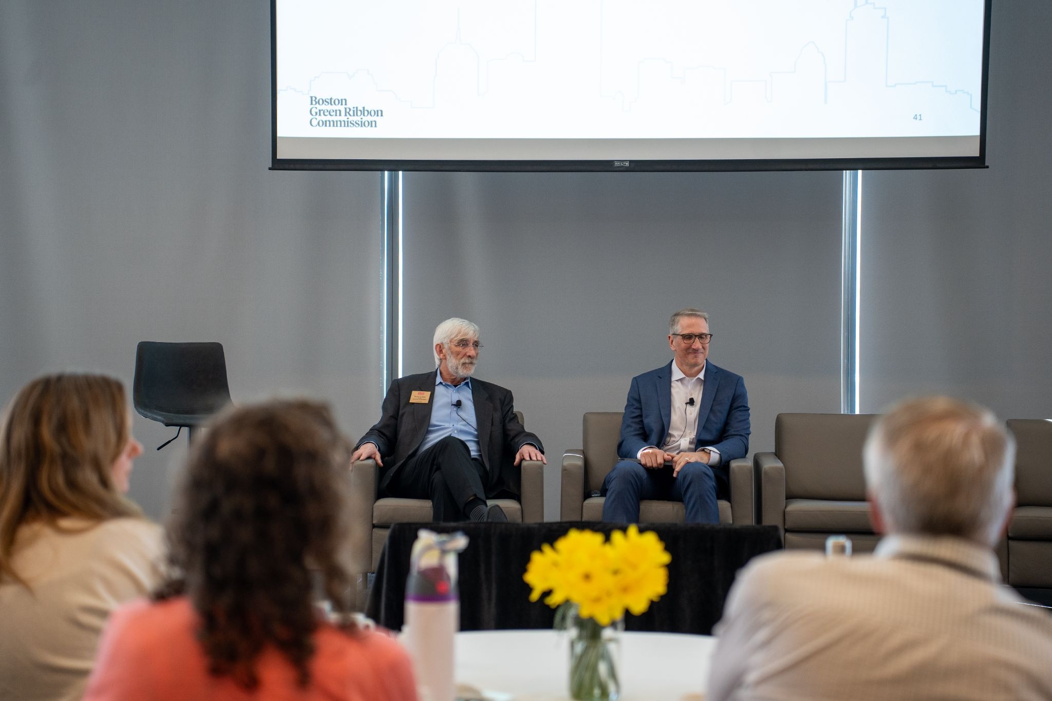 Two men sit on stage in armchairs during a panel discussion, with a projection screen above them. An audience sits facing them; a vase of yellow flowers is in the foreground.