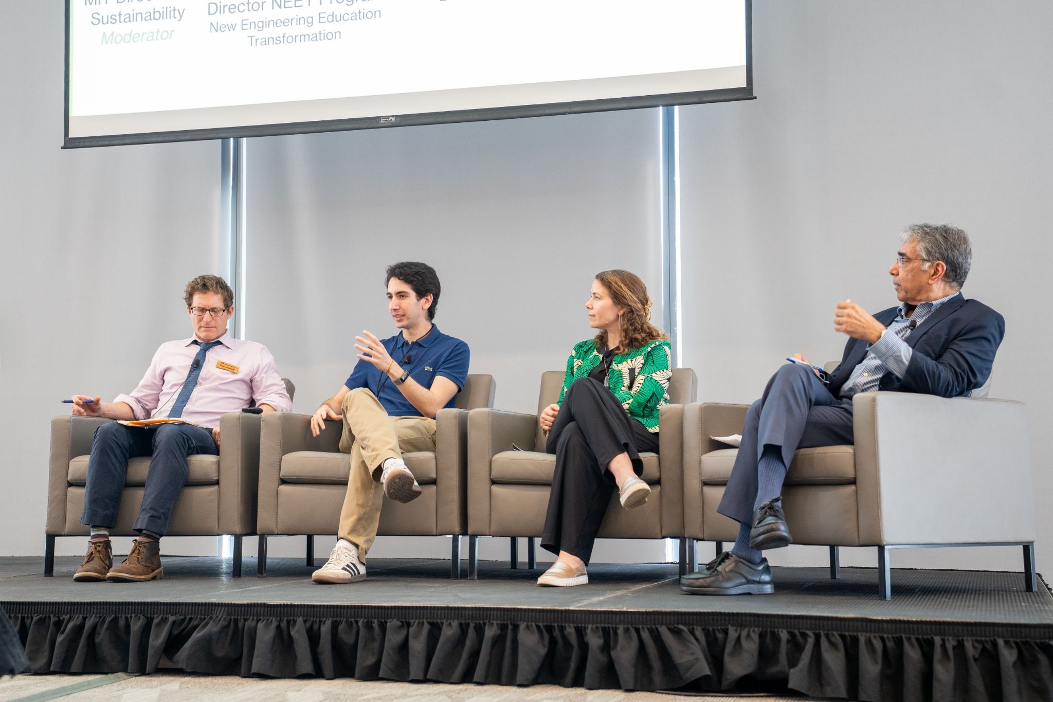 Four panelists sit on stage in armchairs. They are engaged in discussion, appearing thoughtful and attentive. The setting is professional and formal.
