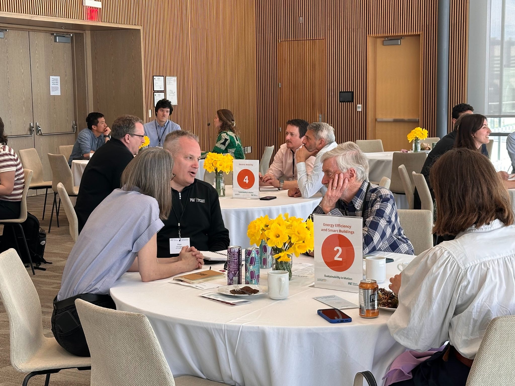 A group of people engaged in discussion around round tables in a conference room, adorned with daffodils. Numbered signs indicate themed topics.