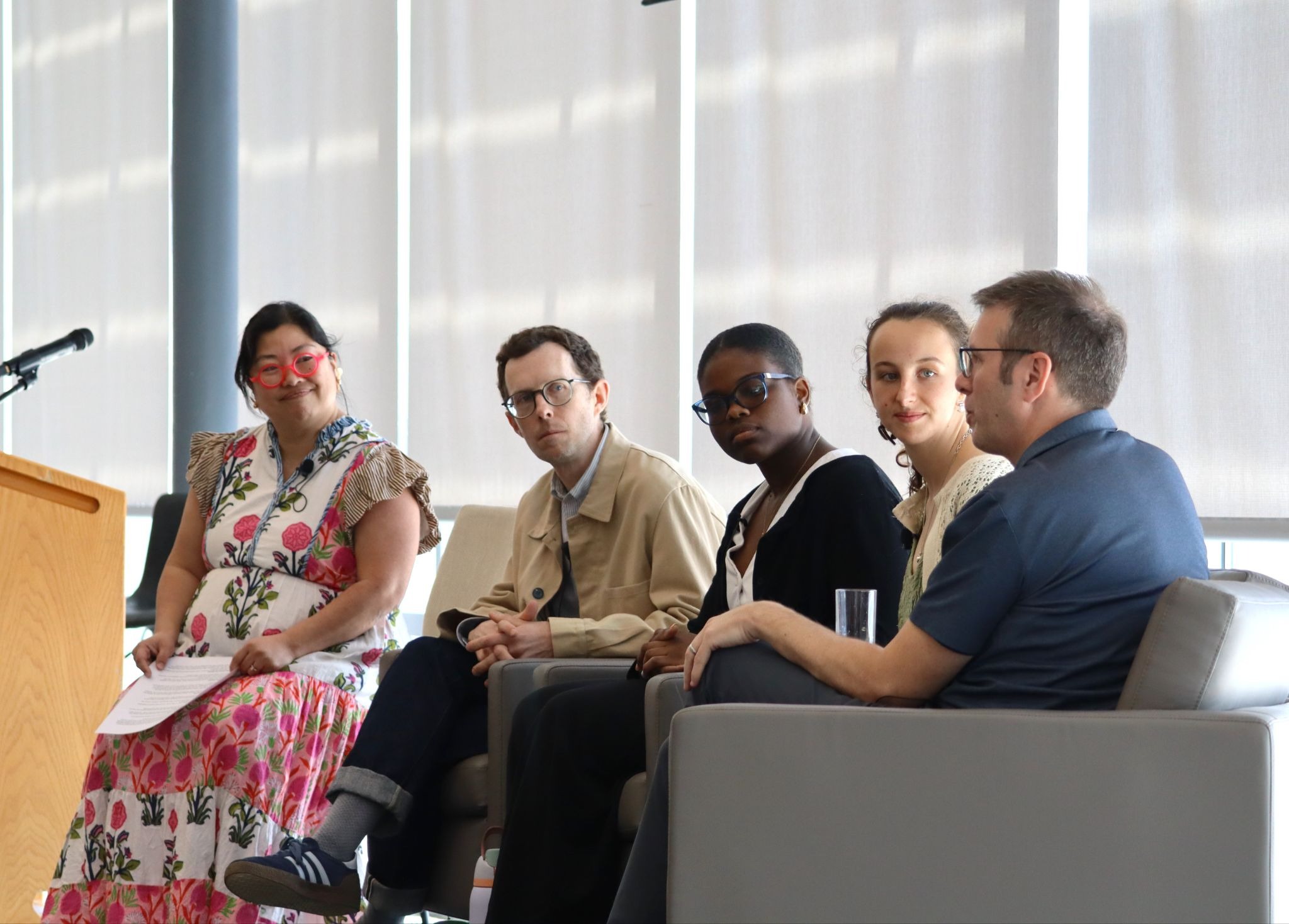 Five people sit in a row of grey armchairs during a panel discussion in a bright, modern room. From left to right: a woman in a floral dress and red glasses, a man in a tan jacket, a person in a black cardigan, a woman in a lace top, and a man in a blue polo who is speaking. They are all looking toward the right of the frame.