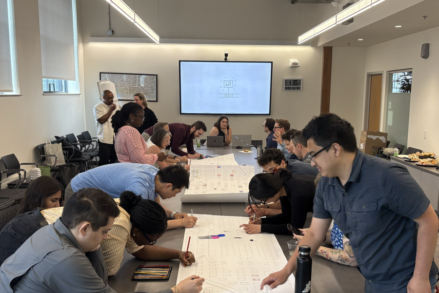A group of MIT students and staff gathered around a conference table are actively engaged in a collaborative activity, writing on large sheets of paper. The room is well-lit, with a presentation screen in the background. The atmosphere is focused and collaborative.