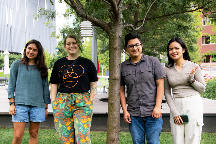 Four students pose together outdoors under a large tree on a campus setting. Above them, a white beehive-shaped heat sensor with a solar radiation shield hangs from one of the tree branches. Modern buildings are visible in the background, with green foliage and grass surrounding the group.