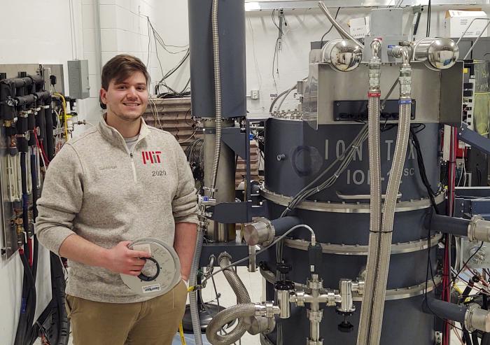 
              MIT graduate student Daniel Korsun holds a reel of the high-temperature superconducting tape that has been the focus of his research, as he stands beside the cyclotron he uses in his experiments. 
              Photo: Steve Jepeal
      