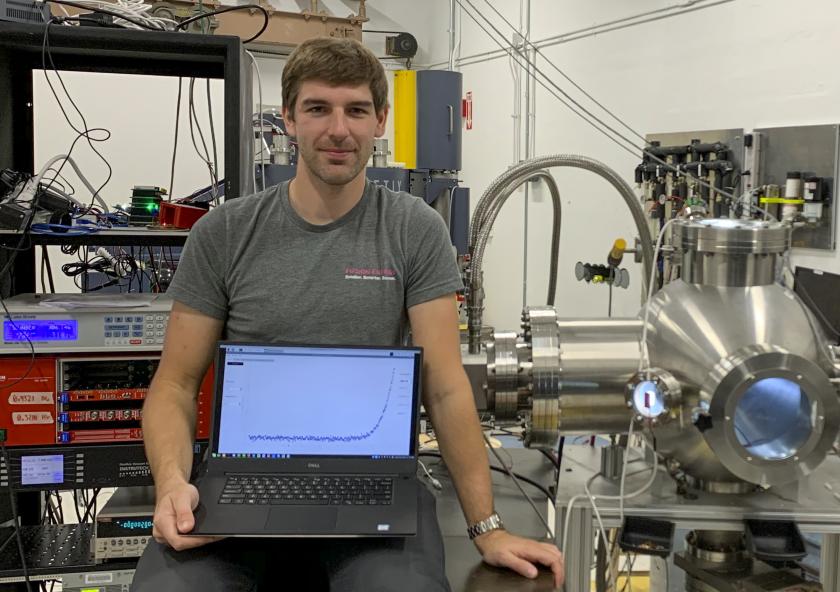 
              David Fischer sits beside the experiment’s vacuum chamber (illuminated in blue), where the high temperature superconductor tapes will be mounted for proton irradiation and in situ transport current measurement. His laptop shows dat...