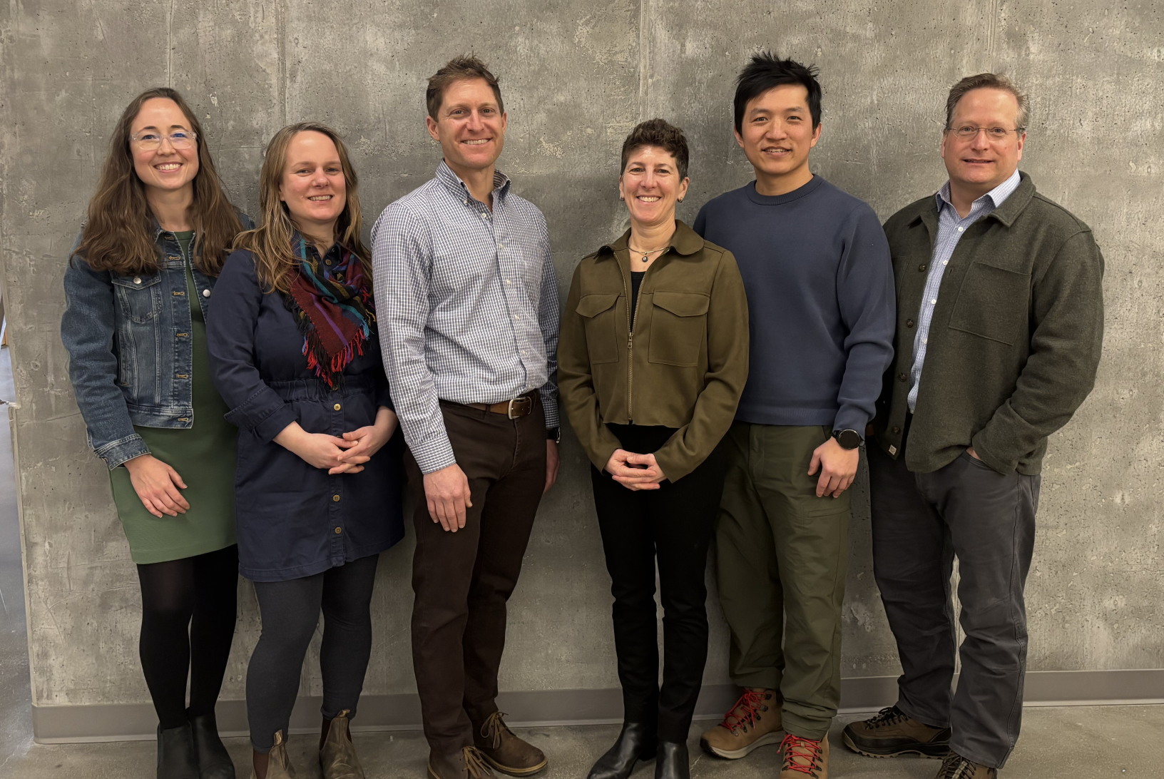 Six people stand smiling in front of a gray wall. They wear casual attire, expressing a friendly and relaxed atmosphere.