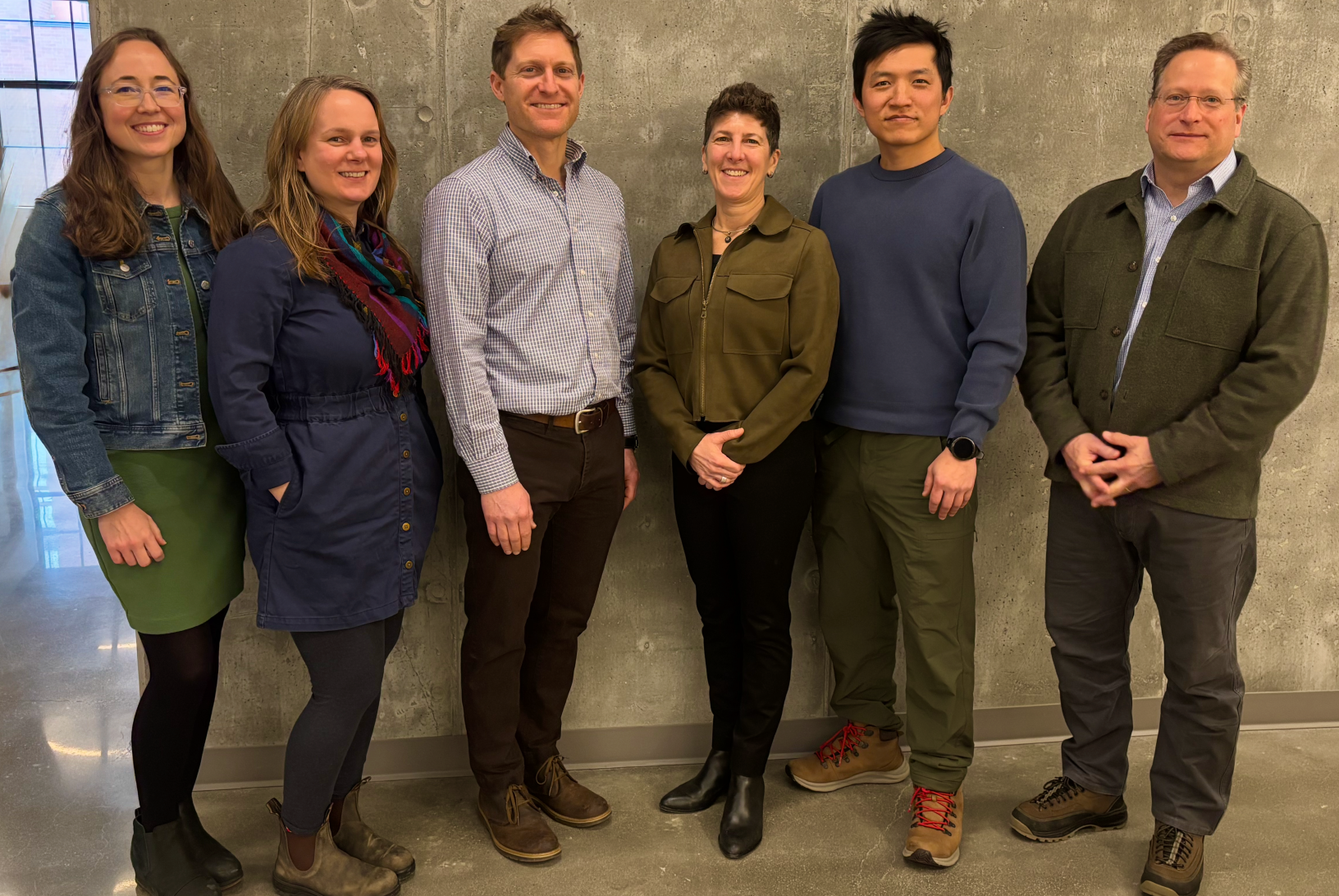 Six people stand smiling in front of a gray wall. They wear casual attire, expressing a friendly and relaxed atmosphere.