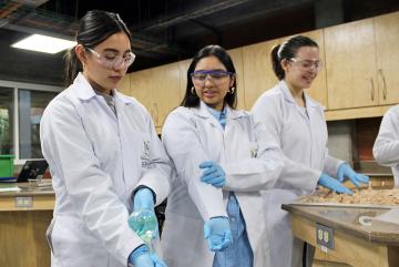 Project Manager Lourdes Figueroa teaches a student how to handle a volumetric flask to prepare one of the chemical solutions used in the reactions for the process. The other students are observing closely as they follow the steps of the demonstration, which is part of the initial stages of chemical preparation for the production of chitosan nanoparticles.