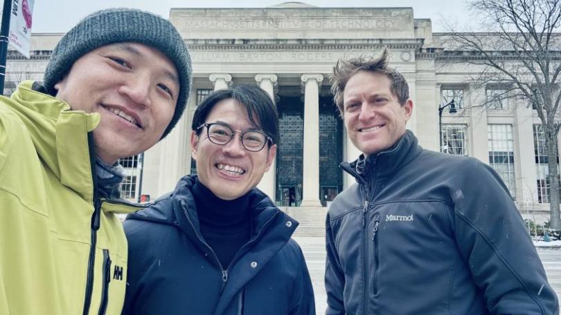 Three people smiling in front of a grand, columned building with "Massachusetts Institute of Technology" inscribed above the entrance. They appear cheerful and bundled warmly, suggesting a cold day.