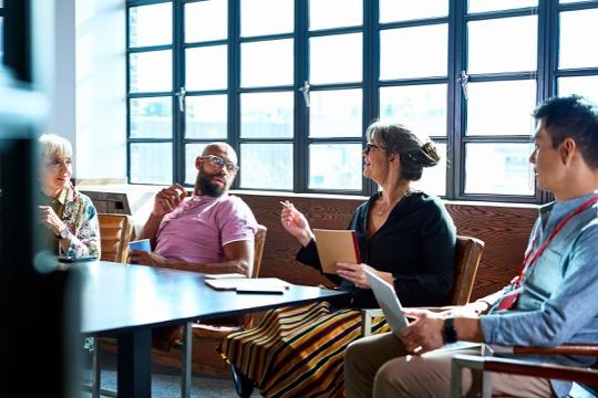 A group of four people engaged in discussion around a table in a well-lit room with large windows. The mood appears collaborative and attentive.